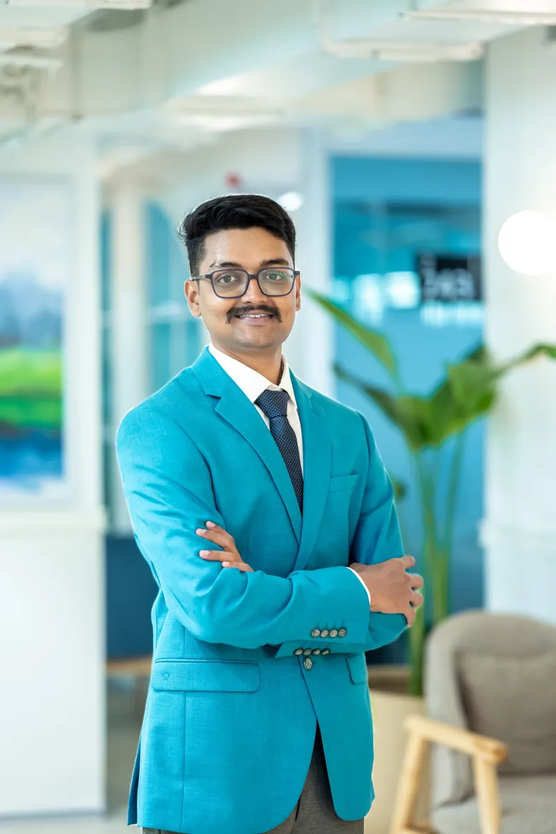 Nachiketh Desai, standing with his arms crossed, in a teal blazer and glasses.