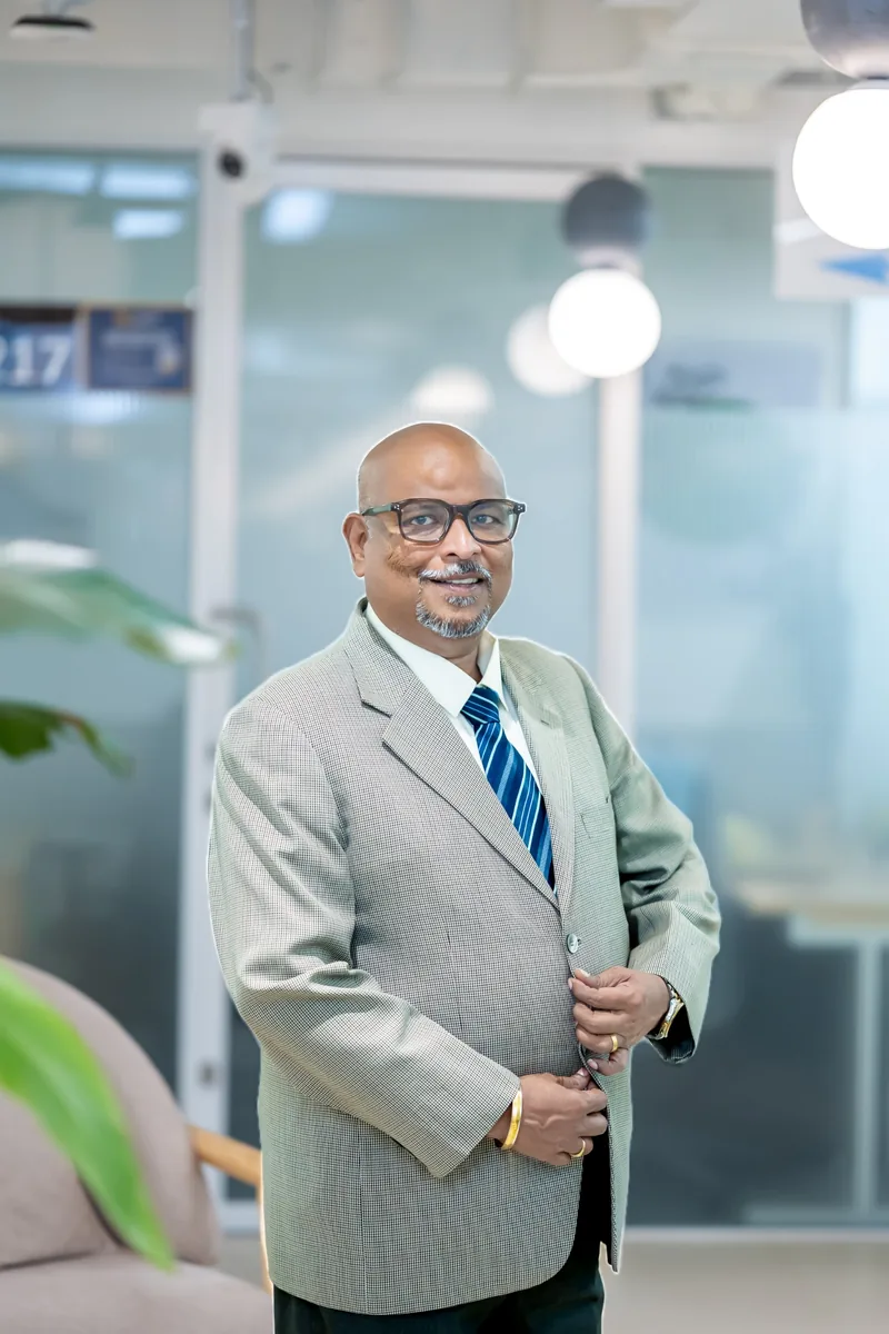 Sudheer Sivapuram, smiling, in a light grey blazer and striped tie.