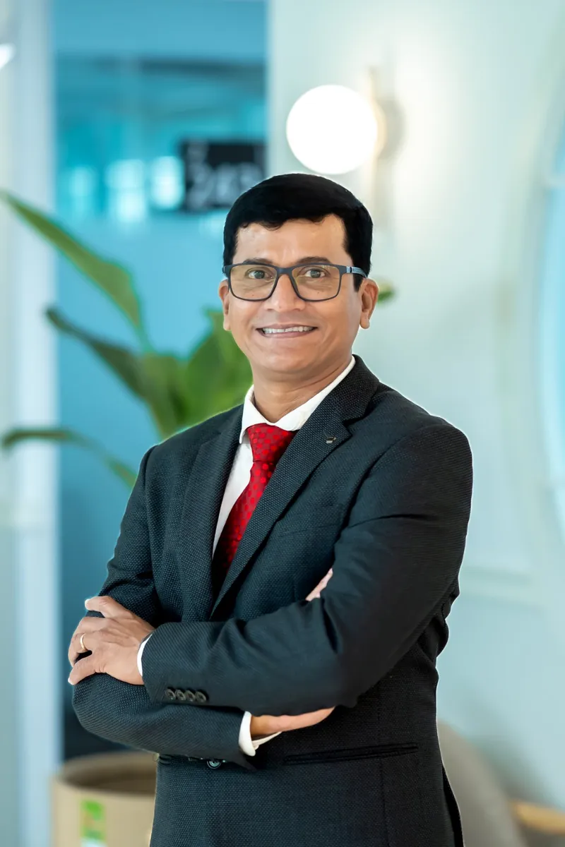Surya Shailendra Kumar, smiling with his arms crossed, in a dark suit and red tie.